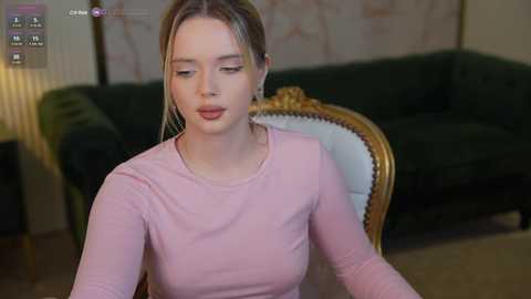 Media: Video of a fair-skinned, blonde-haired woman in a light pink long-sleeve top, seated on a vintage dark green velvet sofa with gold accents. Background shows a blurred, patterned wall.