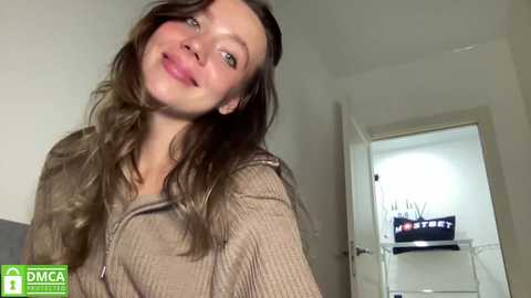 Media: A video of a young woman with long, wavy brown hair, smiling. She's wearing a ribbed, beige sweater, standing indoors near a door with a coat hanger.