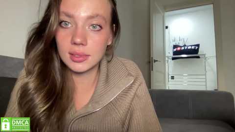 Media: Video of a young woman with fair skin, green eyes, and wavy brown hair, wearing a beige ribbed sweater, sitting indoors.