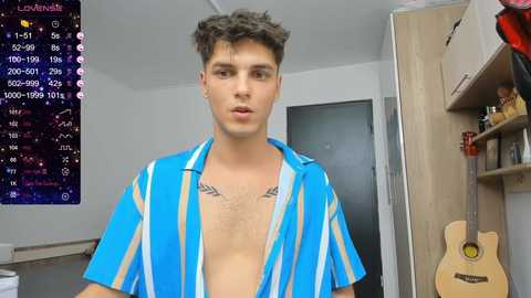 Media: Video of a young, attractive, light-skinned man with short, curly brown hair wearing a blue and white striped shirt unbuttoned to reveal his chest, standing in a cluttered room with a guitar and a music stand.