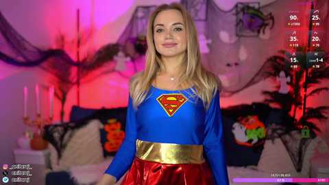 Media: Video of a smiling young woman in a blue Superwoman costume with a red and yellow chest emblem, standing in a Halloween-decorated living room.