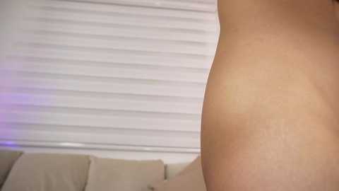 Media: Video of a nude woman's lower torso, showing tan lines on her skin, with a blurred white curtain and beige couch in the background.