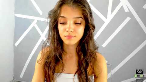 Media: A video of a young girl with long, wavy brown hair, light skin, and a serene expression, wearing a strapless white top. The background features abstract white geometric shapes against a gray wall.