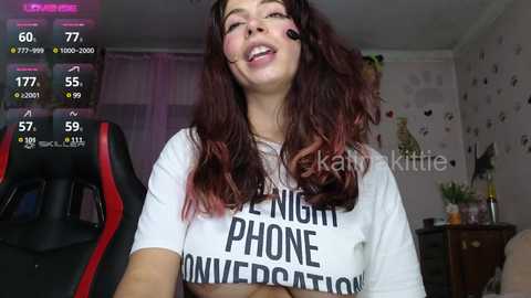 Media: A video of a smiling woman with long, wavy red hair, wearing a white T-shirt with bold text, in a cluttered bedroom with a gaming chair and colorful wall art.