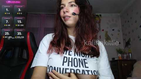 Media: Video of a young woman with long, wavy red hair, wearing a white T-shirt with black text, headset, and gaming chair, in a dimly lit room with gaming overlays.