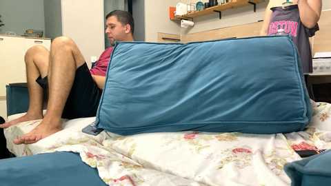 Media: Video of a young man with short dark hair in a hospital bed, wearing a maroon shirt and black shorts, resting on a blue pillow. The room has a blue chair, floral bedspread, and a wooden shelf with medical supplies in the background.