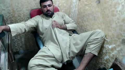 Media: Video of a bearded man in a beige shalwar kameez, lying on a red chair in a rustic room with yellowish walls, looking distressed.