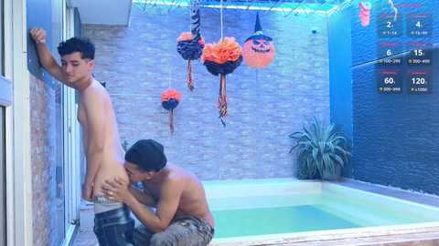 Media: Video of two shirtless men in an indoor pool area. One man is standing nude, leaning against a wall, while the other, wearing a cap, performs oral sex. Halloween decorations and a digital timer are visible.