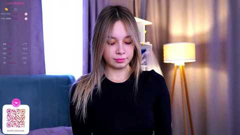 Media: Video of a young woman with straight, shoulder-length blonde hair, wearing a black long-sleeve top, looking down. Background includes a teal couch, a lit floor lamp, and a digital clock.