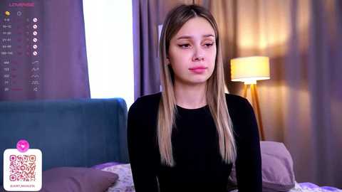 Media: Video of a young Caucasian girl with long, straight blonde hair, wearing a black top, sitting on a bed in a softly lit bedroom.