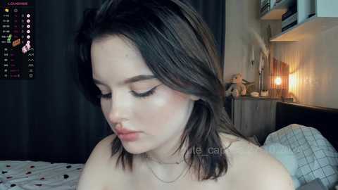 Media: Video of a young woman with shoulder-length dark hair, closed eyes, and a slight smile, wearing a light necklace, in a dimly lit bedroom with a calendar, wall art, and a bed.