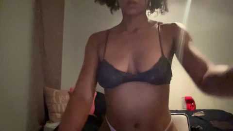 Media: A video of a light-skinned woman with curly hair, wearing a dark gray lace bra and white panties, standing in a dimly lit bedroom with beige walls.