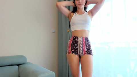 Media: Video of a young woman with light skin and long dark hair, wearing a white crop top and patterned high-waisted shorts, standing in a bright, minimalistic room with a light gray couch and sheer white curtains.