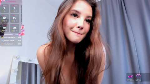 Media: Video of a young woman with long, straight brown hair, wearing a white top, smiling gently. Background features a digital clock and grey curtains in a minimalist room.