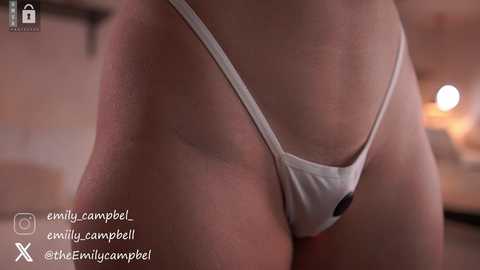 Media: Video of a close-up view of a woman's lower torso in a white thong, showing a hint of pubic hair. The background is blurred, featuring a warm, indoor setting. Text in the bottom left corner reads \"@campbell@thecamperel.\