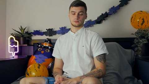 Media: Video of a young man with short brown hair and tattoos, sitting cross-legged on a bed, wearing a white shirt and blue shorts. Background features Halloween decorations, including a glowing pumpkin and orange pillows.