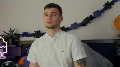 Media: Video of a young man with short dark hair and a beard, wearing a white button-up shirt, standing in a room adorned with Halloween decorations.