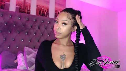 Media: Video of a Black woman with braided hair, wearing a black long-sleeve top, standing in a bedroom with a tufted headboard, cityscape wallpaper, and pink lighting.