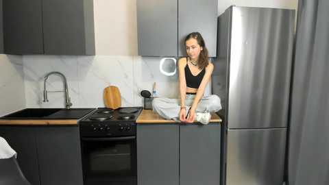 Media: Video of a slender, light-skinned woman with straight black hair, wearing a black tank top and jeans, sitting on a kitchen counter next to a stainless steel fridge in a modern, gray-toned kitchen with marble backsplash.