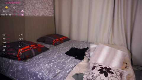 Media: A video of a messy bedroom with two unmade beds covered in gray and floral-patterned bedding. Black clothes are scattered on the bed, and a plush toy rests nearby.