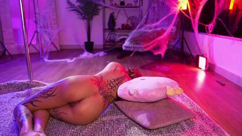 Media: Video of a naked woman with tattoos, lying on a couch in a dimly lit, modern room with purple and pink lights, ferns, and a tent.