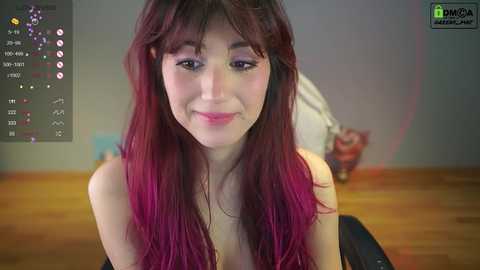 Media: A video of a smiling young woman with long, dark red hair and fair skin, wearing minimal makeup, sitting in a dimly lit room with wooden floors and a cluttered background.