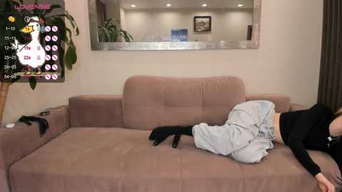 Media: Video of a woman sleeping on a pink sofa, wearing a black top and gray pants, in a living room with a mirror, potted plant, and gaming overlay.