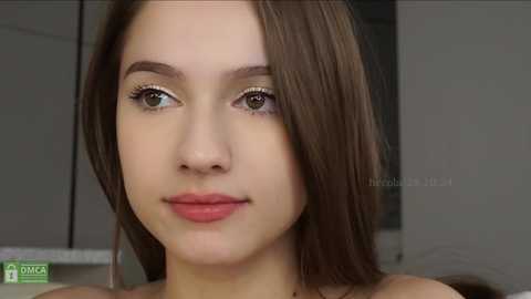 Media: Video of a young Caucasian woman with straight brown hair, light skin, and light makeup, including mascara and light pink lipstick. Background is out of focus, possibly indoor setting.