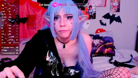 Media: A photo of a pale-skinned woman with blue hair, wearing a black off-shoulder top, black choker, and cat ears, lying on a bed in a room with bat decorations and a pink light.