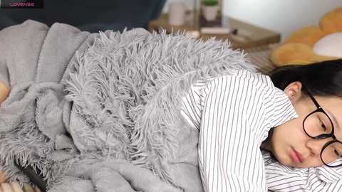 Media: Video of an East Asian woman with long black hair and glasses, wearing a striped shirt, snuggled under a gray, fluffy blanket on a bed.