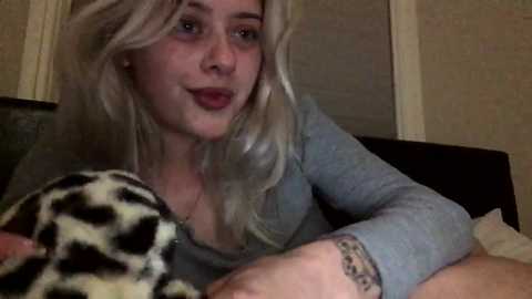 Media: A video of a young blonde woman with light skin, wearing a gray top, smiling while holding a black and white cow-patterned blanket. The background features beige walls and a dark couch.