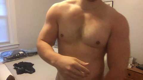 Media: Video of a naked, muscular, light-skinned man with small nipples, standing in a bedroom with a white bed, clothes, and a window in the background.