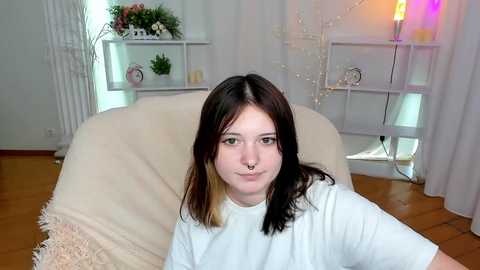 Media: Video of a young woman with pale skin, long dark hair, and a septum piercing, wearing a white T-shirt, seated on a beige couch in a bright, minimalist room with white walls, shelves, and a wooden floor.