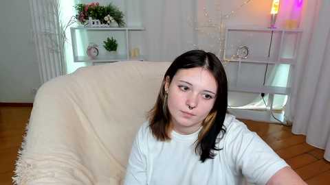 Media: Video of a young Caucasian woman with long black hair, fair skin, and a septum piercing, wearing a white T-shirt, sitting on a cream-colored sofa in a minimalist living room with wooden floors and white decor.