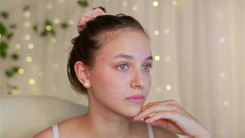 Media: Video of a young, light-skinned Caucasian woman with fair skin and brown hair in a messy bun, wearing a white spaghetti-strap top, posing contemplatively against a softly lit, bokeh background.