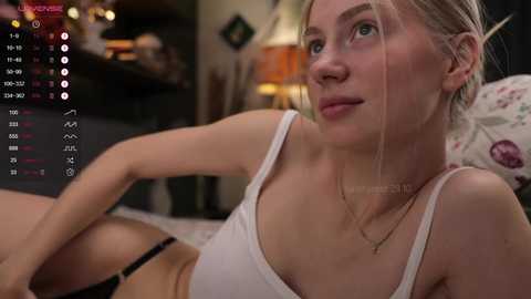 Media: Video of a blonde woman with fair skin, wearing a white tank top, lying on a bed in a dimly lit room, with a virtual reality headset on.