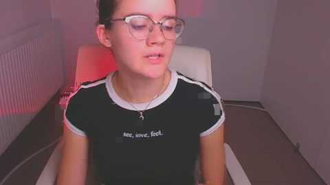 Media: Video of a young woman with light skin, glasses, and dark hair in a black athletic shirt with white trim, seated on a white chair in a dimly lit room with red lighting.