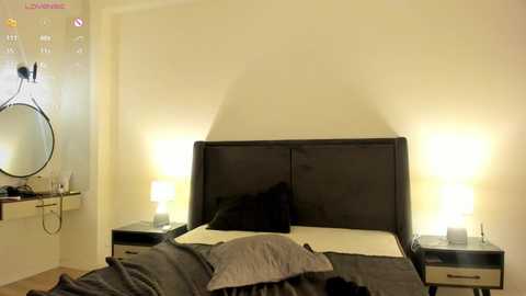 Media: Video of a minimalist, modern bedroom with a dark wood headboard, beige walls, two bedside tables with white lamps, a round mirror, and a dark bed with gray pillows.