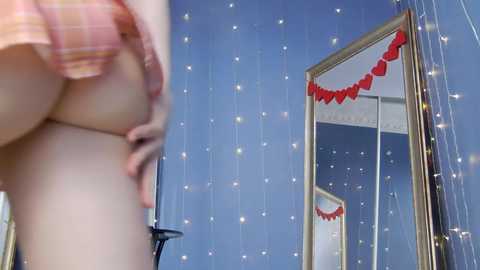 Media: Video of a young woman with fair skin, wearing a pink plaid shirt, lifting her shirt to reveal her large breasts, standing in a room with a mirrored wardrobe and heart garland.