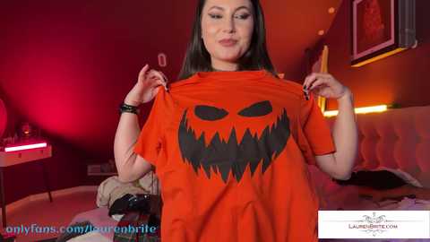 Media: A video of a woman with long dark hair, wearing an orange T-shirt with a large, black jack-o'-lantern design, smiling, in a dimly lit, red-lit room.