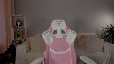 Media: Video of a pink gaming chair with a futuristic cockpit design, placed in a cozy living room with beige couches, a bookshelf, and a potted plant.