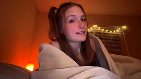 Media: Video of a young Caucasian woman with light skin, brown hair in pigtails, wearing a beige blanket, smiling. Warm, dim orange light fills the room, with string lights and a lit candle in the background.