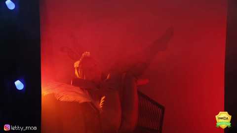 Media: Video of a woman in a dark, smoky room with red lighting, lying on a bed, legs raised, wearing a yellow dress, with the watermark \"Iggy moa\" on the left.