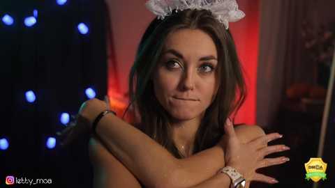 Media: Video of a smiling woman with long brown hair, wearing a white lace headband and bracelets, hugging herself in a dimly lit room with blue and red lights.