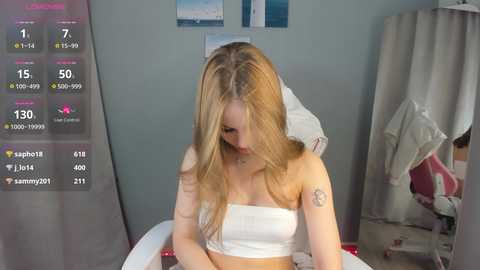 Media: Video of a young blonde woman in a white strapless top, sitting on a bed in a messy room.