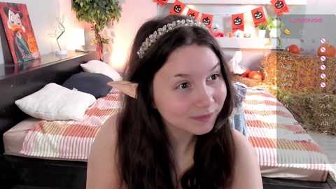 Media: Video of a young woman with long brown hair, wearing a princess crown and elf ears, smiling in a brightly decorated, cozy bedroom with a bed, plant, and Halloween decor.