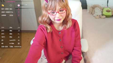 Media: Video of a fair-skinned woman with shoulder-length wavy hair and red glasses, wearing a red cardigan, sitting on a white chair in a bedroom with a wooden floor and teddy bears on the bed.