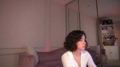 Media: Video of a woman with shoulder-length, wavy black hair, wearing a white robe, standing in a minimalist, softly lit living room with beige walls, a purple couch, and a white bookshelf.