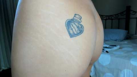 Media: A video shows a light-skinned person's upper back with a tattoo of a blue bottle cap near their shoulder blade. The background features a bed with white polka-dotted sheets and a black metal headboard.