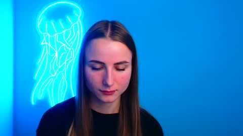 Media: Video of a young woman with long brown hair, wearing a black top, standing against a bright blue wall with a neon jellyfish light.